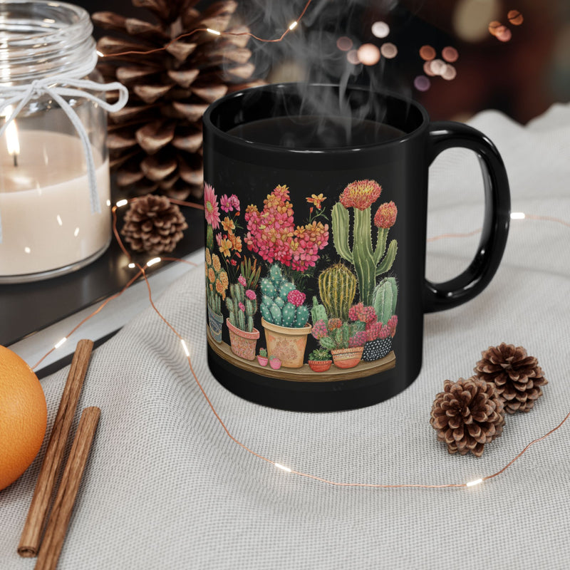 An 11 oz black ceramic mug featuring a vibrant wrap-around cactus garden design, placed on a cozy holiday-themed setup with pinecones, candle, and twinkling lights