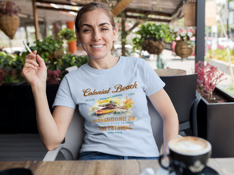 Woman relaxing and smiling at a sunny outdoor cafe wearing a light blue Colonial Beach Premium T-Shirt Playground on the Potomac, holding a cigarette and enjoying her coffee