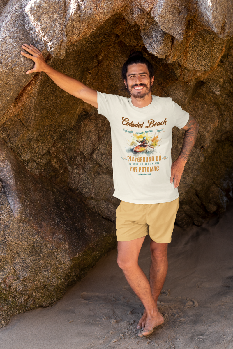 Man leaning casually against a large rocky cave, barefoot on the sand, smiling and wearing the natural color Colonial Beach Premium T-Shirt Playground on the Potomac