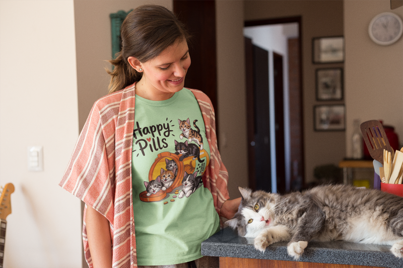 A cheerful woman wearing a mint green "Happy Pills" t-shirt with playful kitten graphics, gently petting her long-haired cat resting on a kitchen counter.