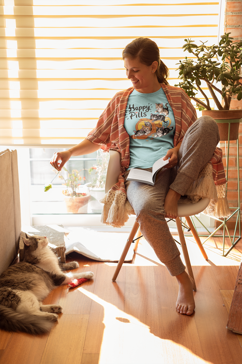 A relaxed woman in a striped shawl sitting by a sunny window wearing a turquoise "Happy Pills" t-shirt with cartoon cats, smiling while playing with her fluffy cat on the floor.