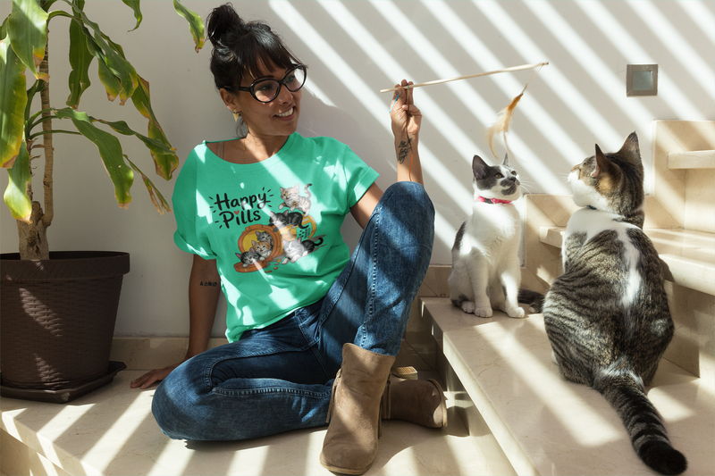 A happy woman wearing glasses and a heather sea green "Happy Pills" t-shirt with adorable cartoon kittens, playing with two curious cats on sunlit indoor stairs.