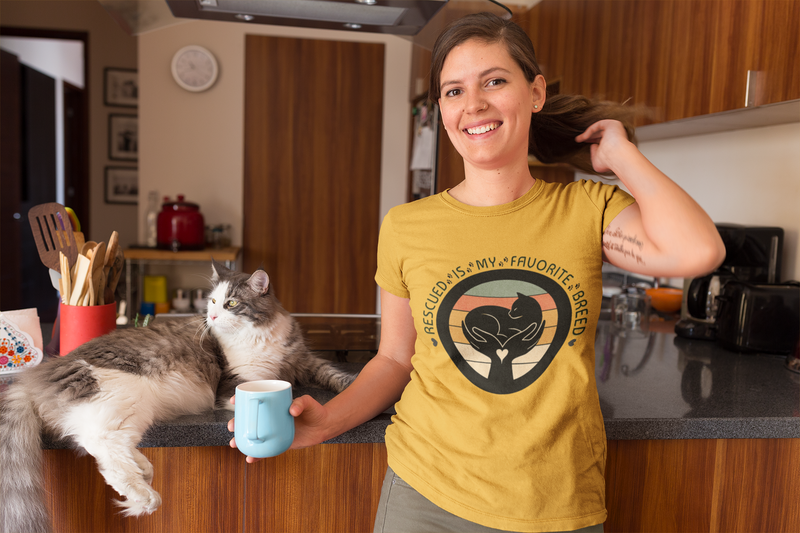 Woman in a mustard yellow t-shirt with a cat graphic in a kitchen holding a mug.