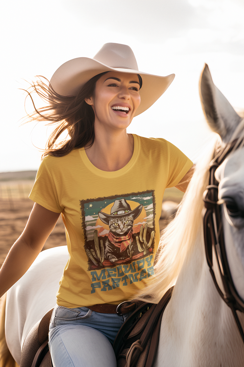 Laughing female model riding a horse wearing the Mustard Yellow Meowdy Partner Western Cat T-Shirt, showcasing a cowboy cat with desert-themed design