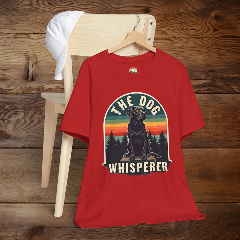 Vibrant red T-shirt featuring a retro design of a black Labrador sitting on a wooden platform, with silhouetted trees and sunset stripes in the background. Text above and below reads “The Dog Whisperer.” Displayed hanging on a wooden chair with a white cap draped on the back.