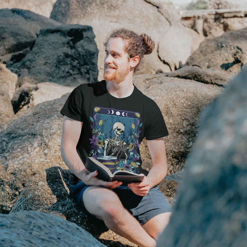 A smiling man sitting on rocks outdoors, wearing The Reader T-Shirt with a mystical gothic tarot card design of a skeleton reading a book, surrounded by colorful flowers and celestial moons on black fabric