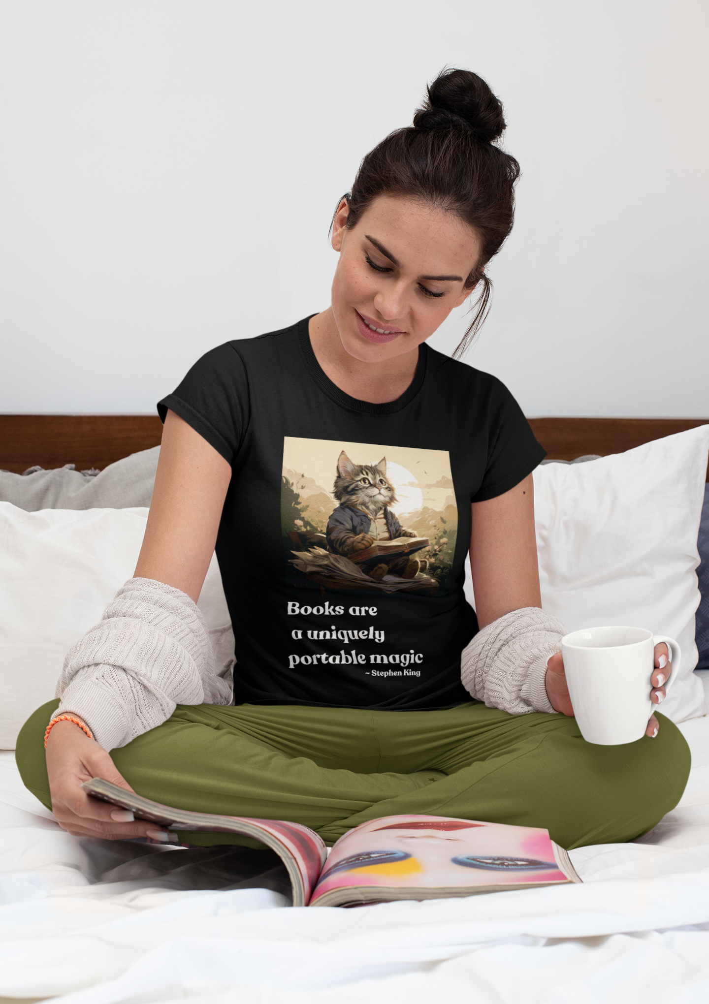 Woman sitting cross-legged on a bed wearing the Books Are a Uniquely Portable Magic cat t-shirt in black.