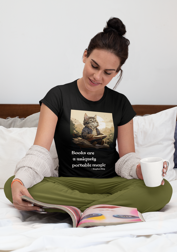 Woman sitting cross-legged on a bed wearing the Books Are a Uniquely Portable Magic cat t-shirt in black.