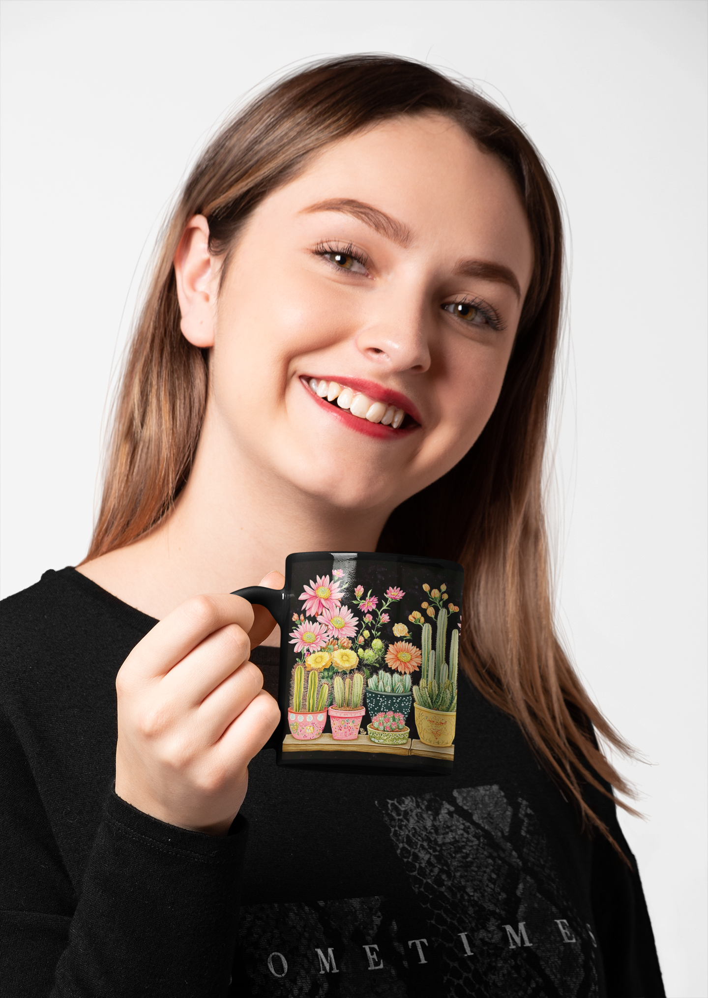 A joyful woman with red lipstick holding an 11 oz black ceramic mug decorated with vibrant blooming cacti in colorful pots, smiling against a white background