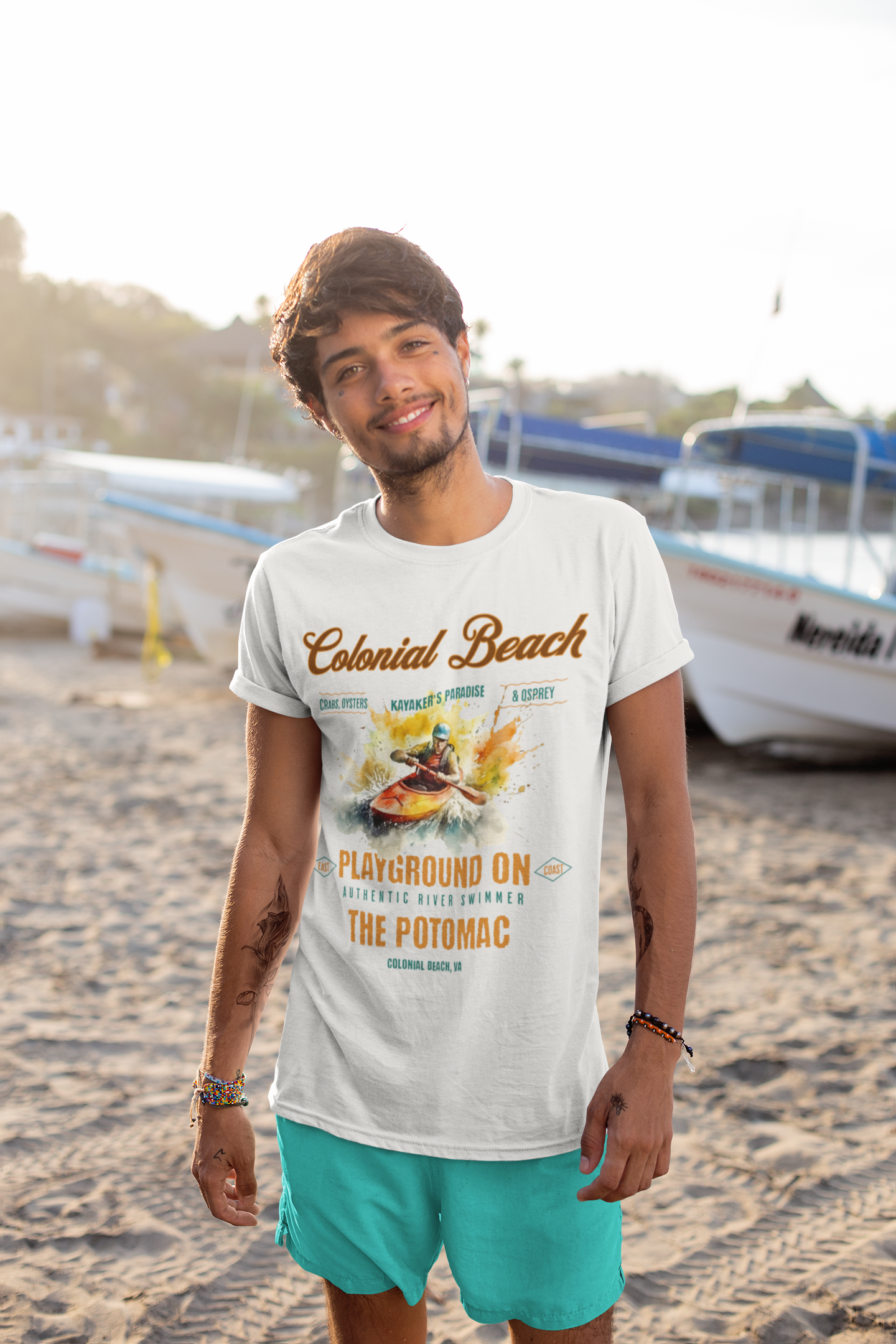 Young man smiling brightly while standing on a sandy beach wearing the ash colored Colonial Beach Premium T-Shirt Playground on the Potomac design, with boats and sunlight in the background