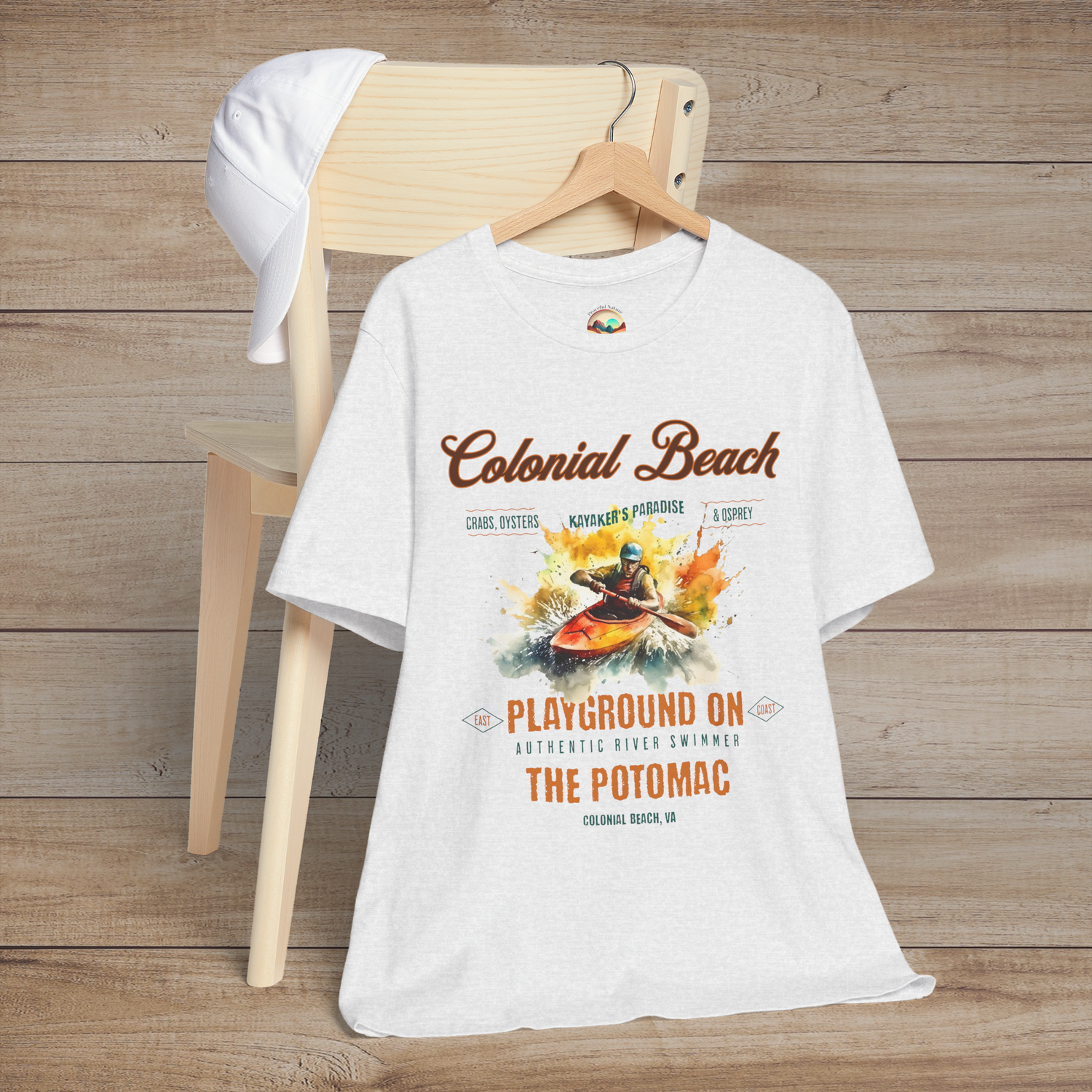 Colonial Beach Premium T-Shirt Playground on the Potomac design displayed on an ash gray t-shirt, laid flat on a wooden chair with a white cap for a relaxed beach vibe