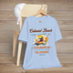 Colonial Beach Premium T-Shirt Playground on the Potomac design neatly arranged on a baby blue t-shirt, hanging on a wooden chair with a casual white cap