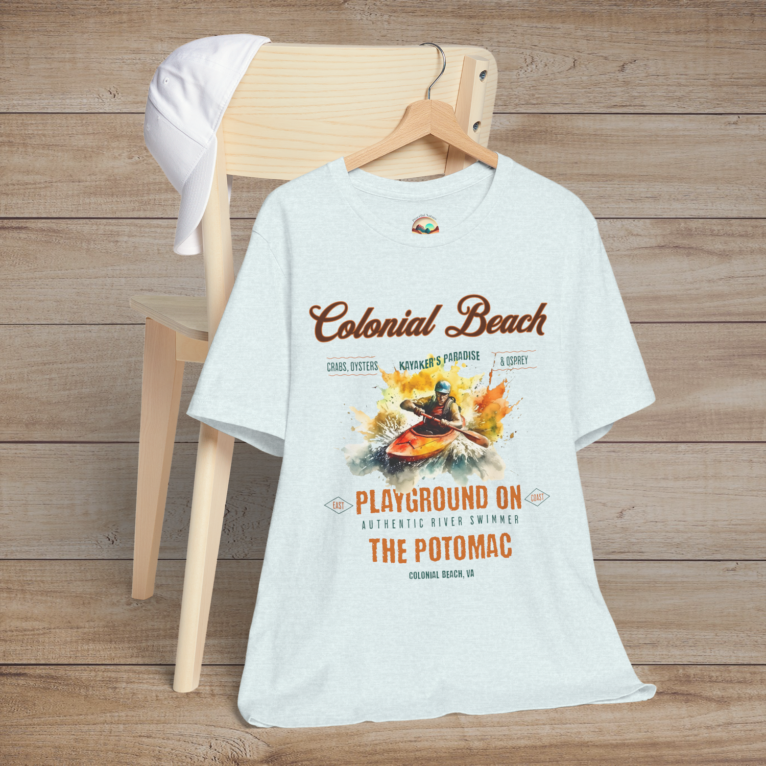 Colonial Beach Premium T-Shirt Playground on the Potomac design laid out on a heather ice blue t-shirt, resting on a chair with a white cap, perfect for outdoor and kayaking fans