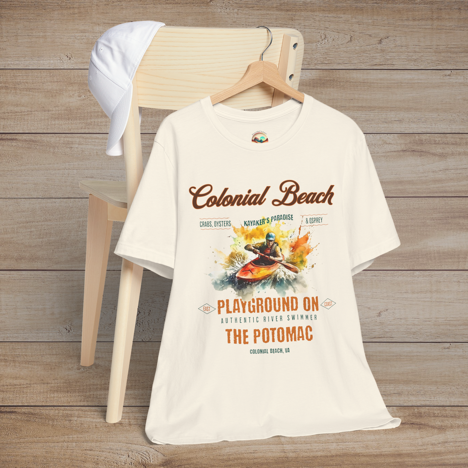 Colonial Beach Premium T-Shirt Playground on the Potomac design featured on a natural color t-shirt, styled against a wood backdrop with a casual cap for coastal aesthetics