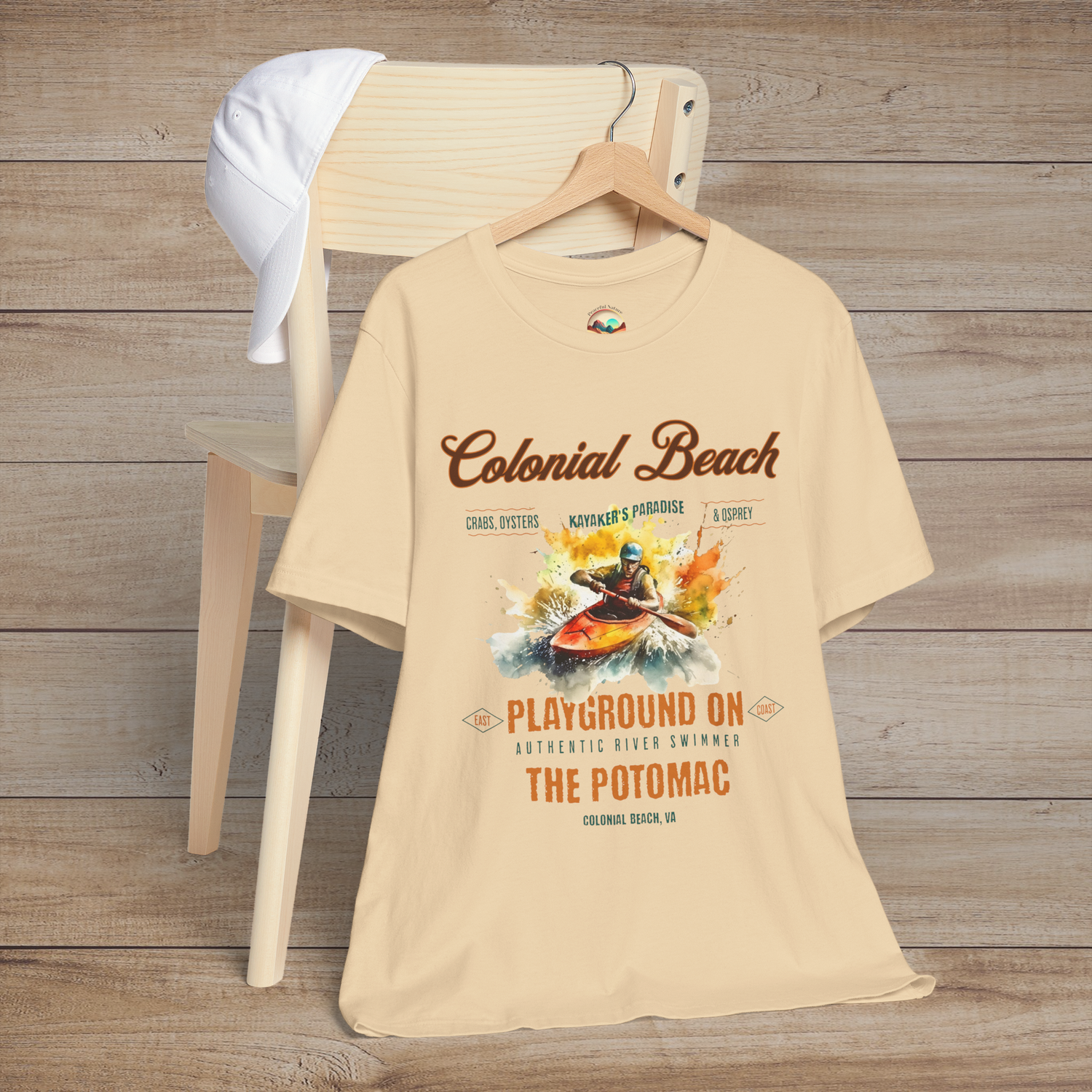 Colonial Beach Premium T-Shirt Playground on the Potomac design printed on a soft cream t-shirt, styled flat on a rustic chair with a cap, evoking beach adventure spirit