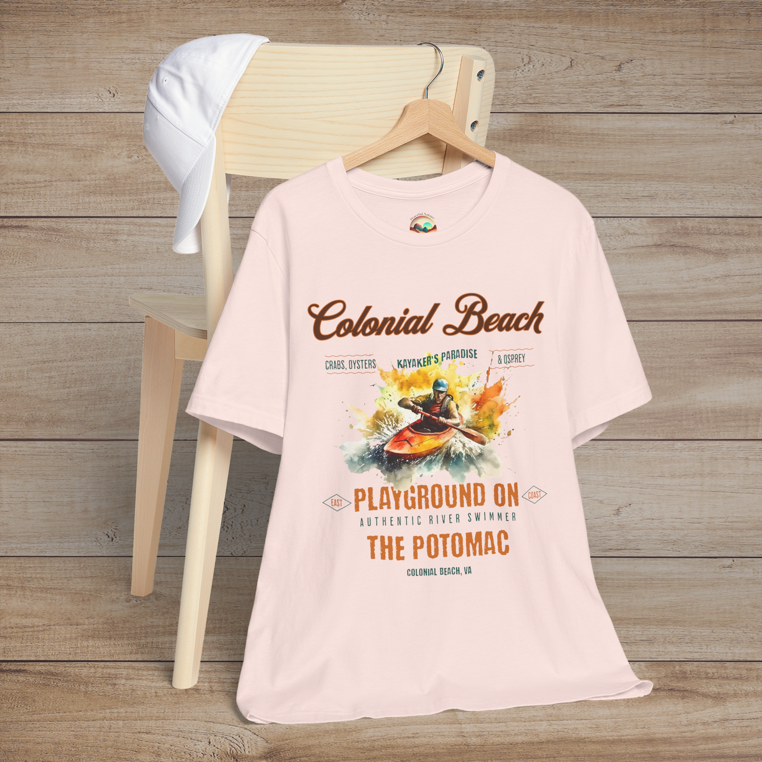 Colonial Beach Premium T-Shirt Playground on the Potomac design showcased on a soft pink t-shirt, placed on a wooden chair next to a white cap, ideal for beach lovers