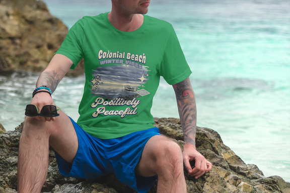 Tattooed man wearing Irish green Colonial Beach Winter Vibes t-shirt sitting on rocks by the ocean