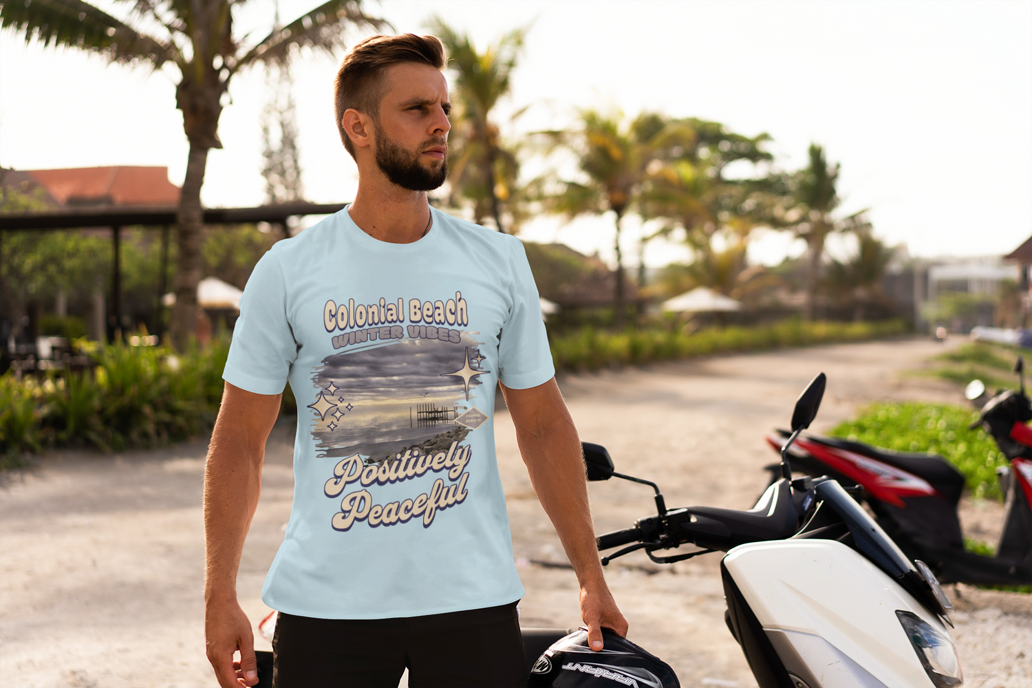 Man wearing light blue Colonial Beach Winter Vibes t-shirt standing next to a motorcycle at the beach under palm trees