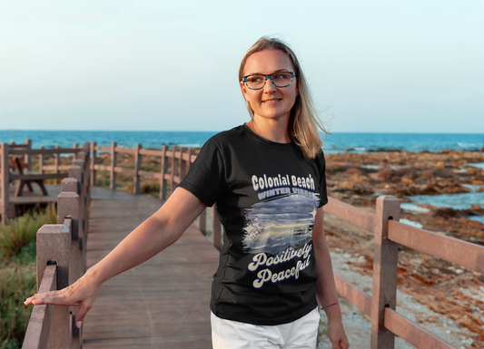 Woman wearing the Colonial Beach Winter Vibes Premium T-Shirt in black while walking on a wooden boardwalk by the rocky ocean shore