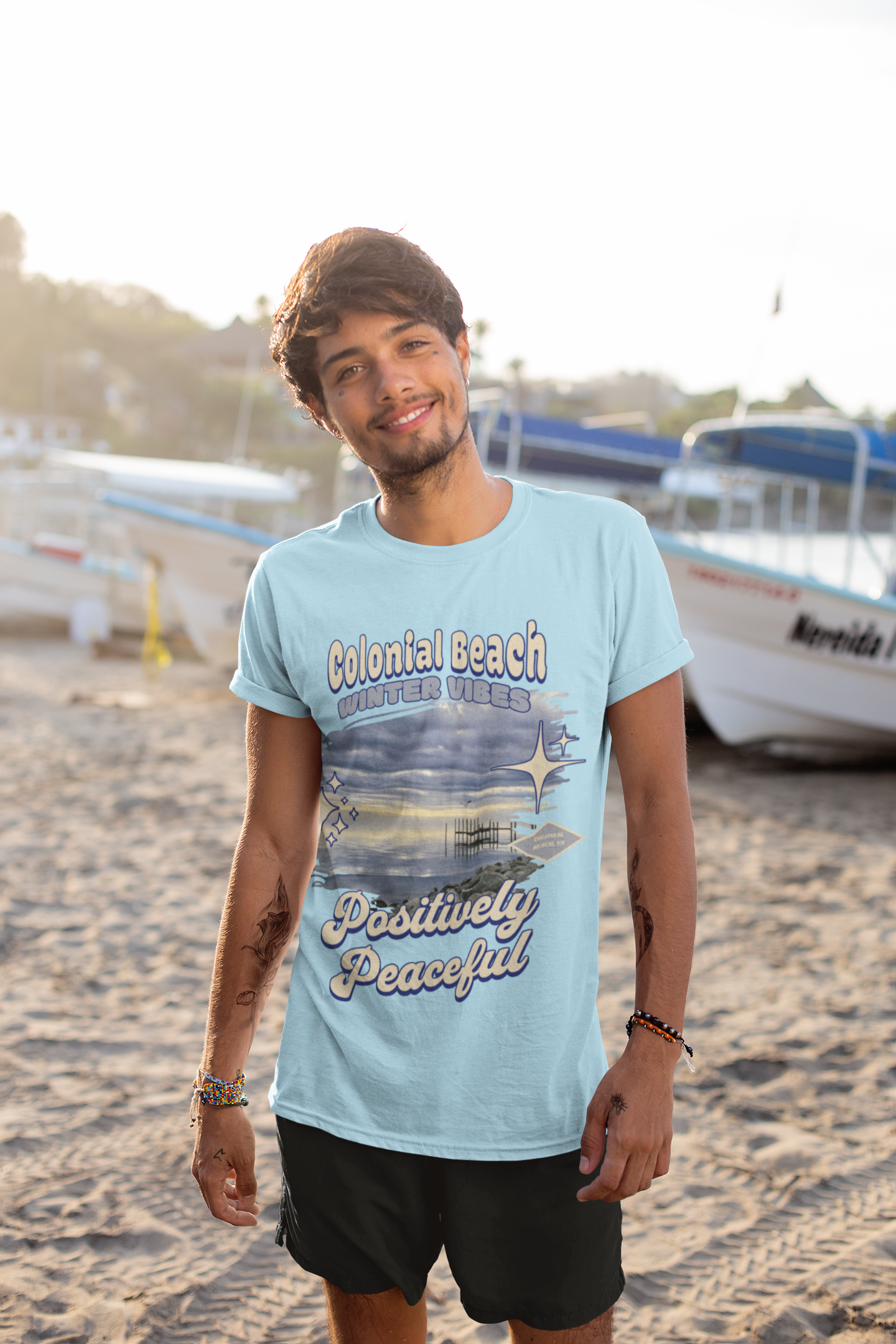 Man wearing Colonial Beach Winter Vibes light blue t-shirt smiling on a beach with boats