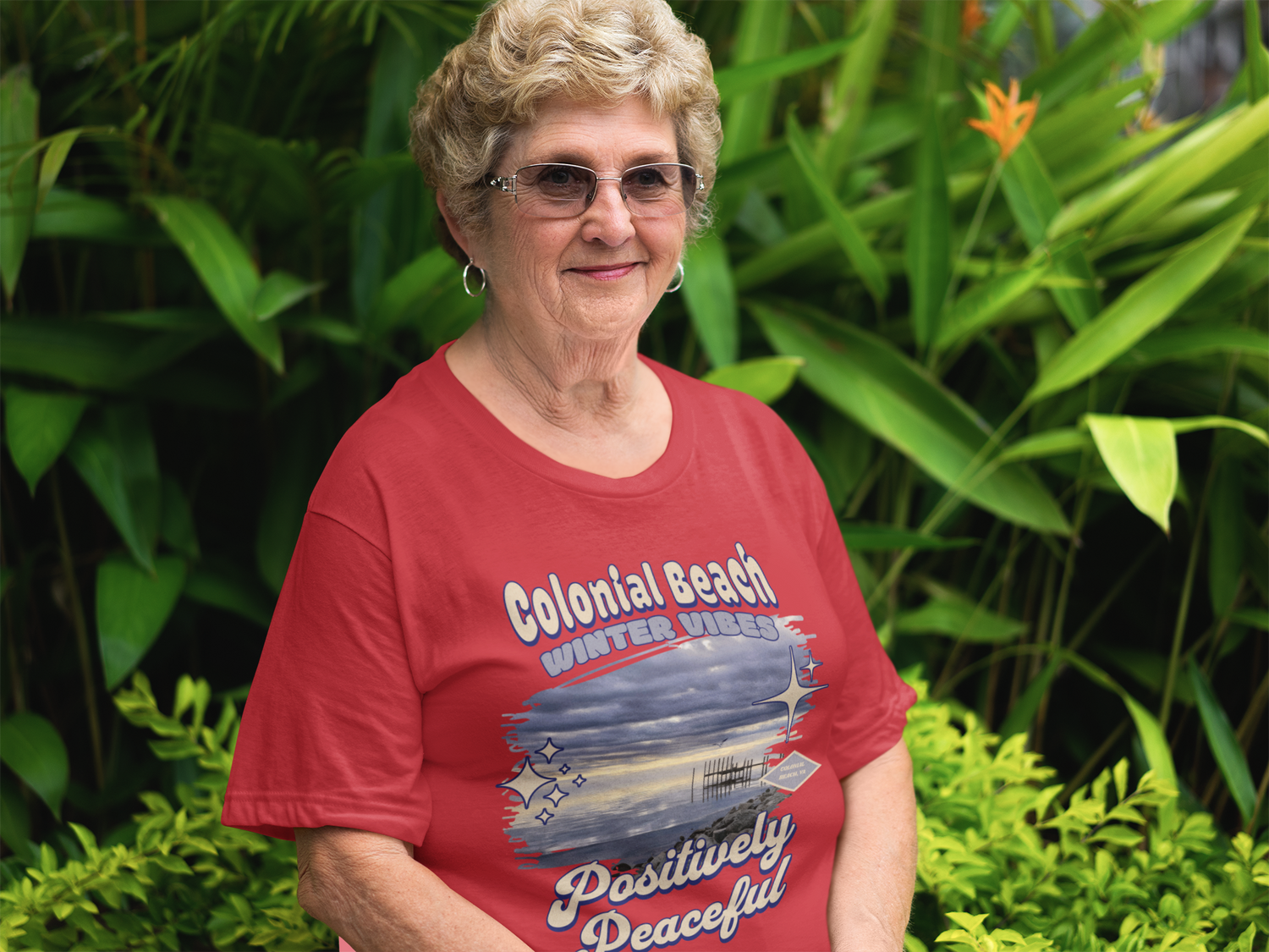 Senior woman smiling while wearing Colonial Beach Winter Vibes red t-shirt in a garden