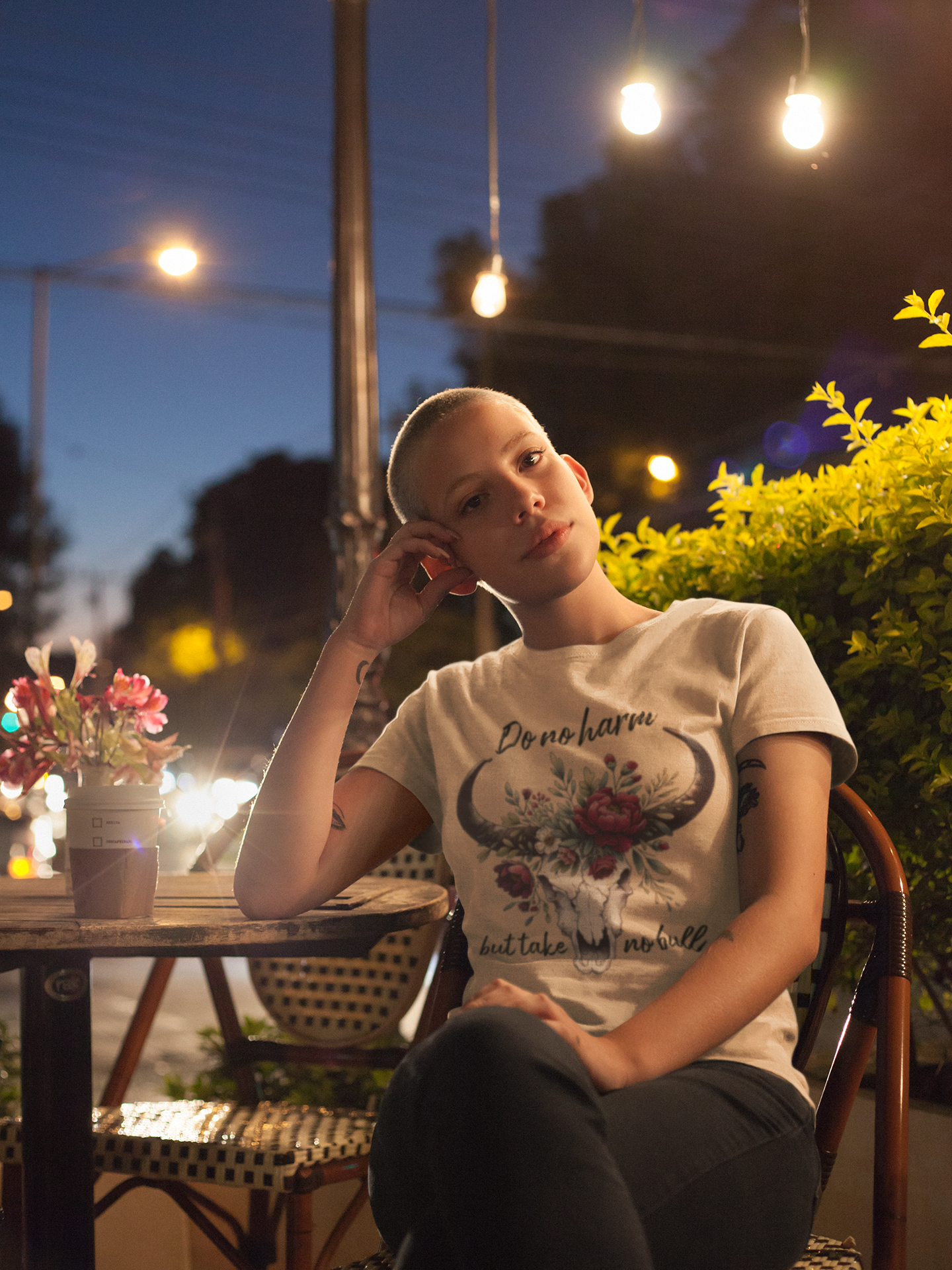 Woman wearing "Do No Harm But Take No Bull" t-shirt in soft cream while sitting at an outdoor café at night