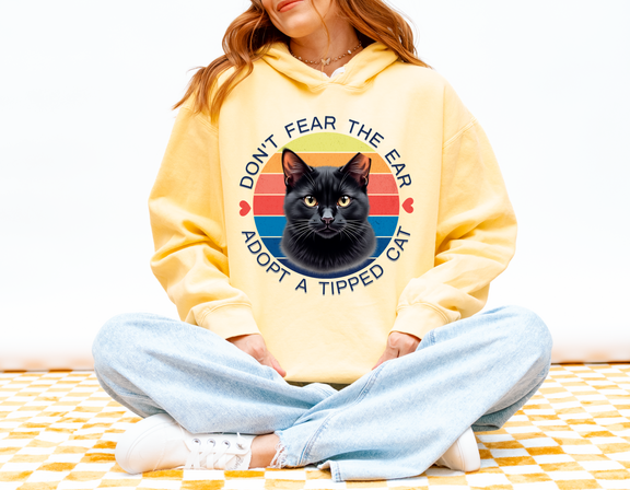 Woman modeling a butter-colored “Don’t Fear the Ear – Adopt a Tipped Cat” hoodie with a vintage-style black cat graphic.