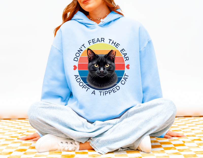 Woman wearing a hydrangea blue hoodie with the “Don’t Fear the Ear – Adopt a Tipped Cat” black cat rescue design.