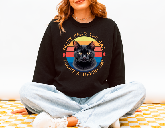 Woman sitting cross-legged wearing a black Gildan 18000 sweatshirt with a retro-style “Don’t Fear the Ear – Adopt a Tipped Cat” black cat design, promoting TNR awareness
