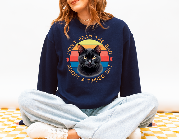 Woman sitting cross-legged wearing a navy blue Gildan 18000 sweatshirt with a retro-style “Don’t Fear the Ear – Adopt a Tipped Cat” black cat design, promoting TNR awareness
