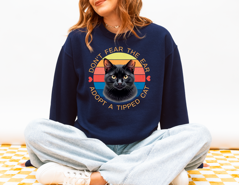 Woman sitting cross-legged wearing a navy blue Gildan 18000 sweatshirt with a retro-style “Don’t Fear the Ear – Adopt a Tipped Cat” black cat design, promoting TNR awareness