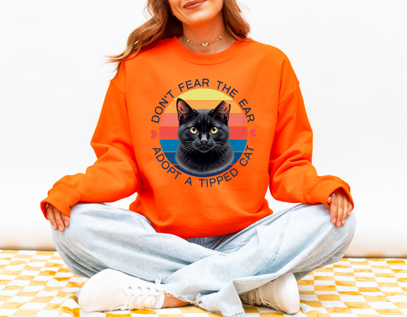 Woman sitting cross-legged wearing a safety orange Gildan 18000 sweatshirt with a retro-style “Don’t Fear the Ear – Adopt a Tipped Cat” black cat design, promoting TNR awareness