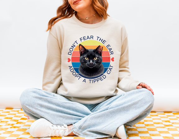 Woman sitting cross-legged wearing a sand-colored Gildan 18000 sweatshirt with a retro-style “Don’t Fear the Ear – Adopt a Tipped Cat” black cat design, promoting TNR awareness