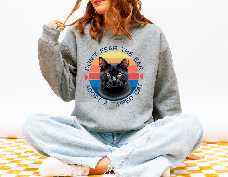 Woman sitting cross-legged wearing a athletic heather (heather grey) Gildan 18000 sweatshirt with a retro-style “Don’t Fear the Ear – Adopt a Tipped Cat” black cat design, promoting TNR awareness