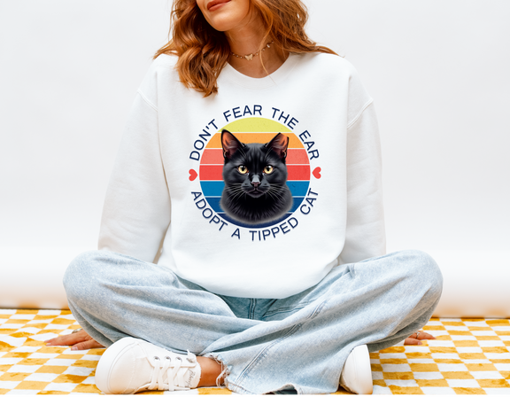 Woman sitting cross-legged wearing a white Gildan 18000 sweatshirt with a retro-style “Don’t Fear the Ear – Adopt a Tipped Cat” black cat design, promoting TNR awareness