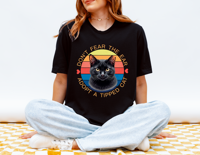 Woman sitting cross-legged wearing a black Bella Canvas 3001 T-shirt with a retro-style “Don’t Fear the Ear – Adopt a Tipped Cat” black cat design, promoting TNR awareness