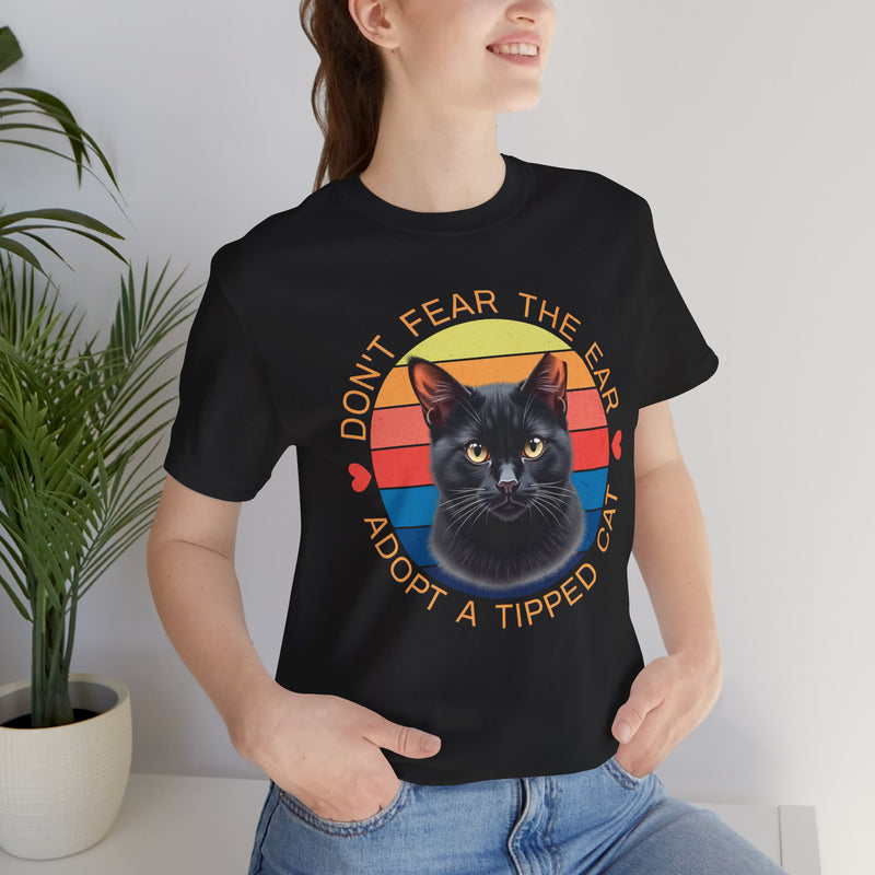 Black Bella Canvas 3001 t-shirt with ear-tipped black cat artwork and retro sunset circle, worn by smiling woman — ‘Don’t Fear The Ear Adopt a Tipped Cat’ feral cat rescue tee