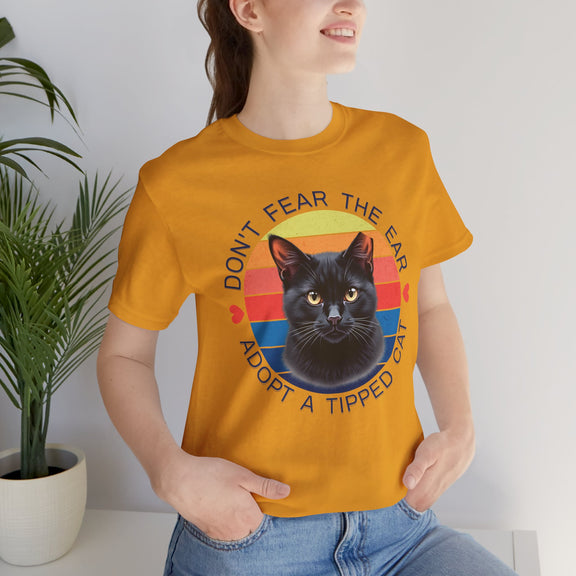 Mustard yellow Bella Canvas 3001 t-shirt showing an ear-tipped black cat graphic with colorful retro stripes, modeled by smiling woman — TNR awareness and adoption advocacy shirt