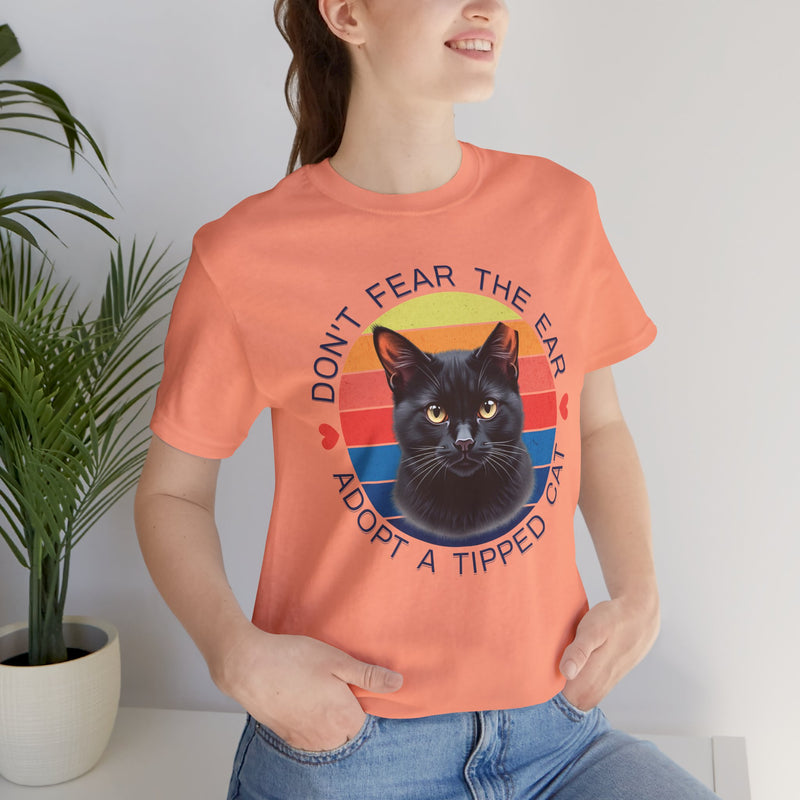 Sunset peach Bella Canvas 3001 t-shirt with ear-tipped black cat illustration and colorful retro circle, worn by woman — TNR cat rescue supporter shirt
