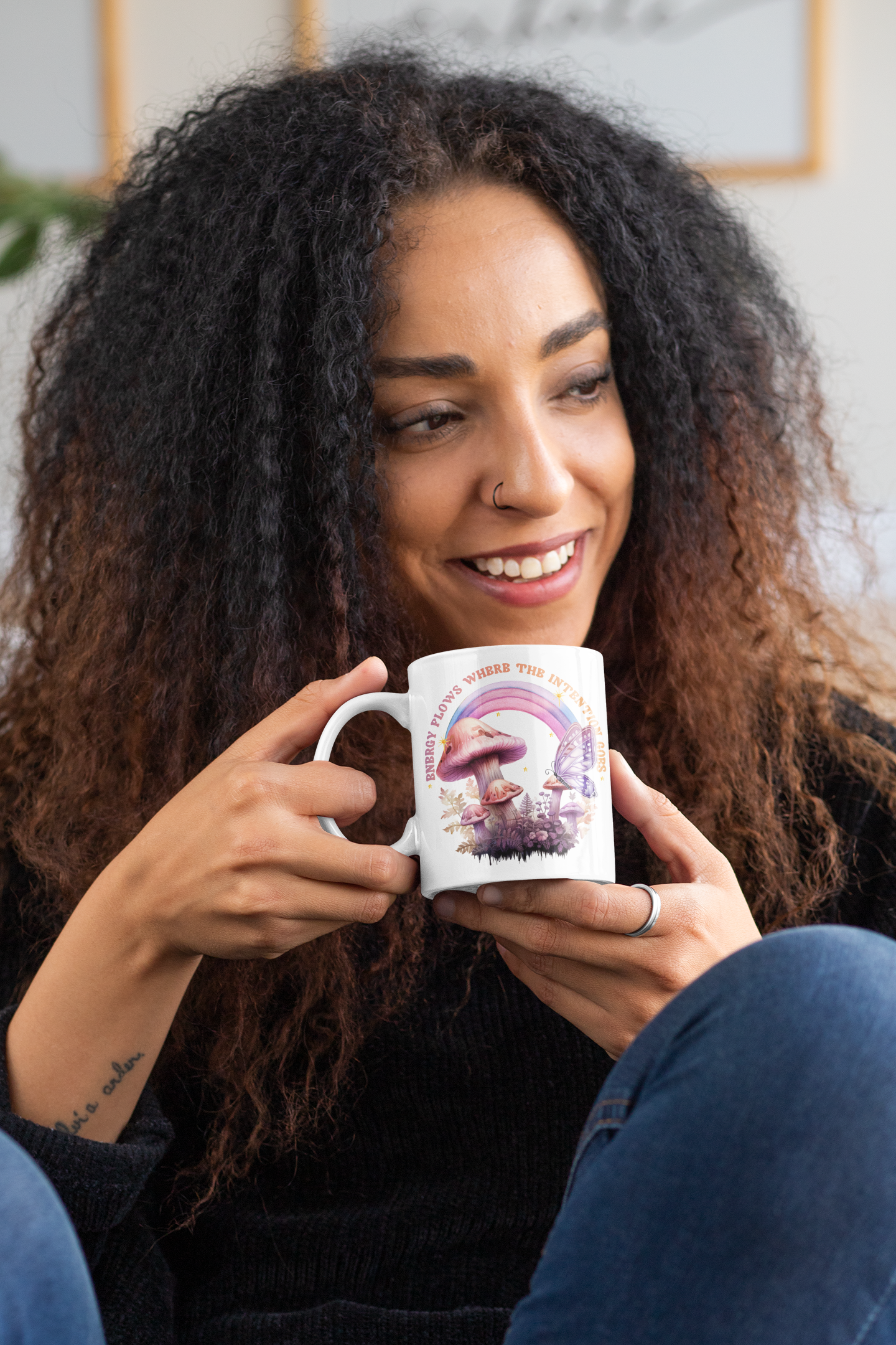 Smiling woman holding an 11 oz white ceramic mug featuring pastel mushrooms, a purple butterfly, and a rainbow with the phrase “Energy flows where the intention goes”