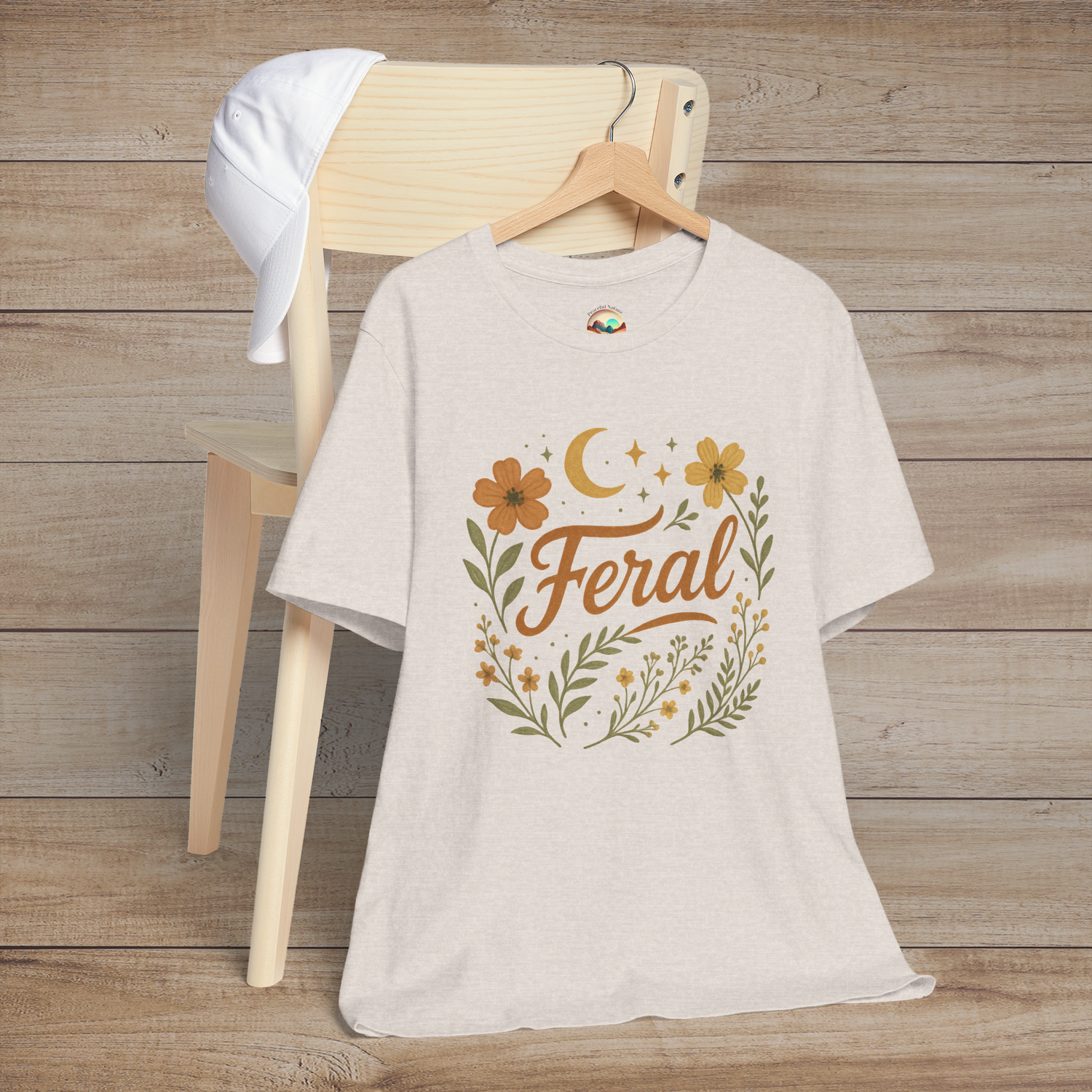 Feral t-shirt in Heather Dust with floral and moon design in elegant calligraphy, displayed on a chair with a cap