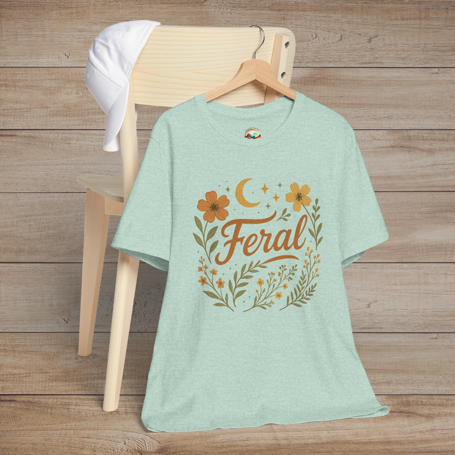 Feral t-shirt in Heather Prism Mint with floral and moon design in elegant calligraphy, displayed on a chair with a cap