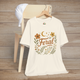 Feral t-shirt in Natural with floral and moon design in elegant calligraphy, displayed on a chair with a cap