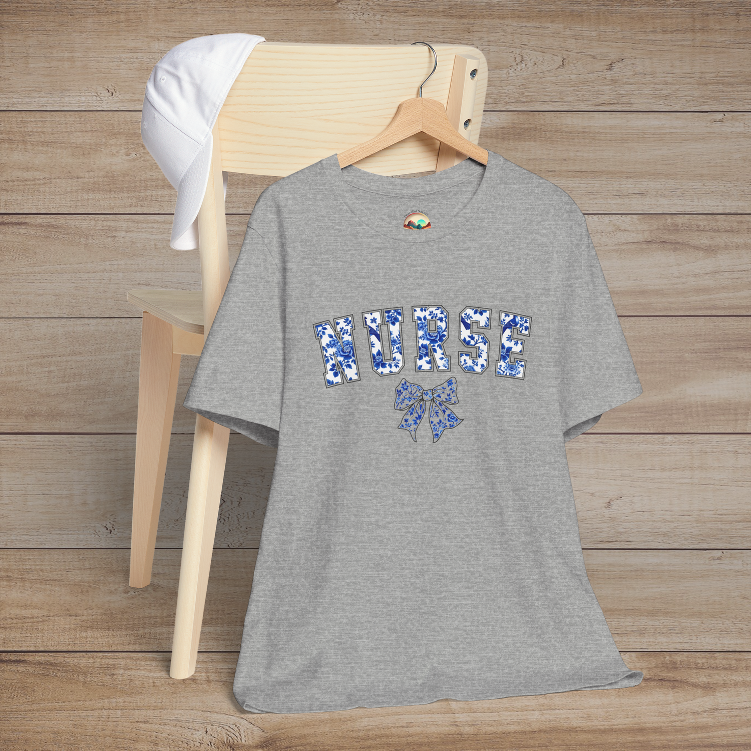 Heather grey t-shirt hanging on a wooden chair, featuring the word 'NURSE' in floral blue letters with a matching bow design – a stylish and elegant tribute to nurses