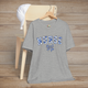 Heather grey t-shirt hanging on a wooden chair, featuring the word 'NURSE' in floral blue letters with a matching bow design – a stylish and elegant tribute to nurses