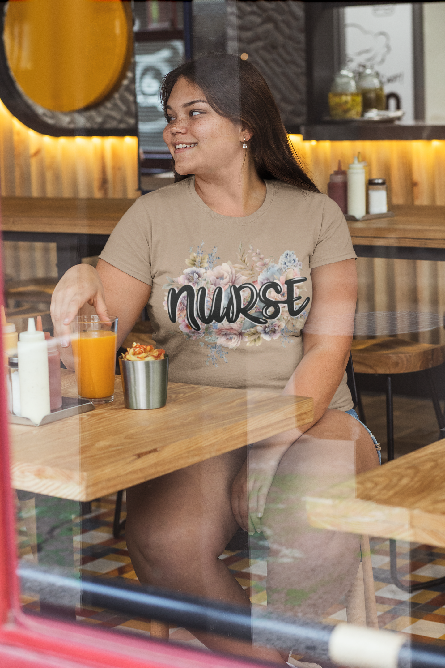 Happy woman at a café modeling the heather tan Nurse Floral Premium Fit T-Shirt, designed with gentle pastel florals and stylish black nurse lettering