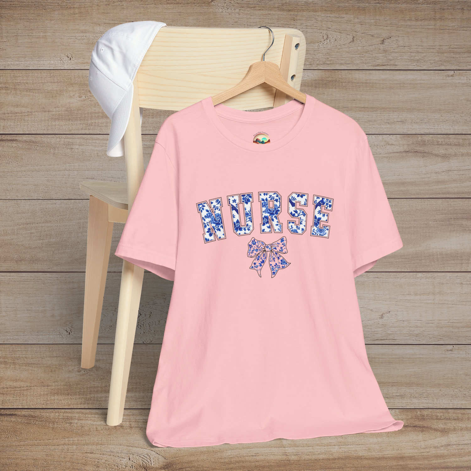 Light pink t-shirt with feminine floral 'NURSE' typography and matching bow, photographed on a hanger – perfect for nurses with a chic style