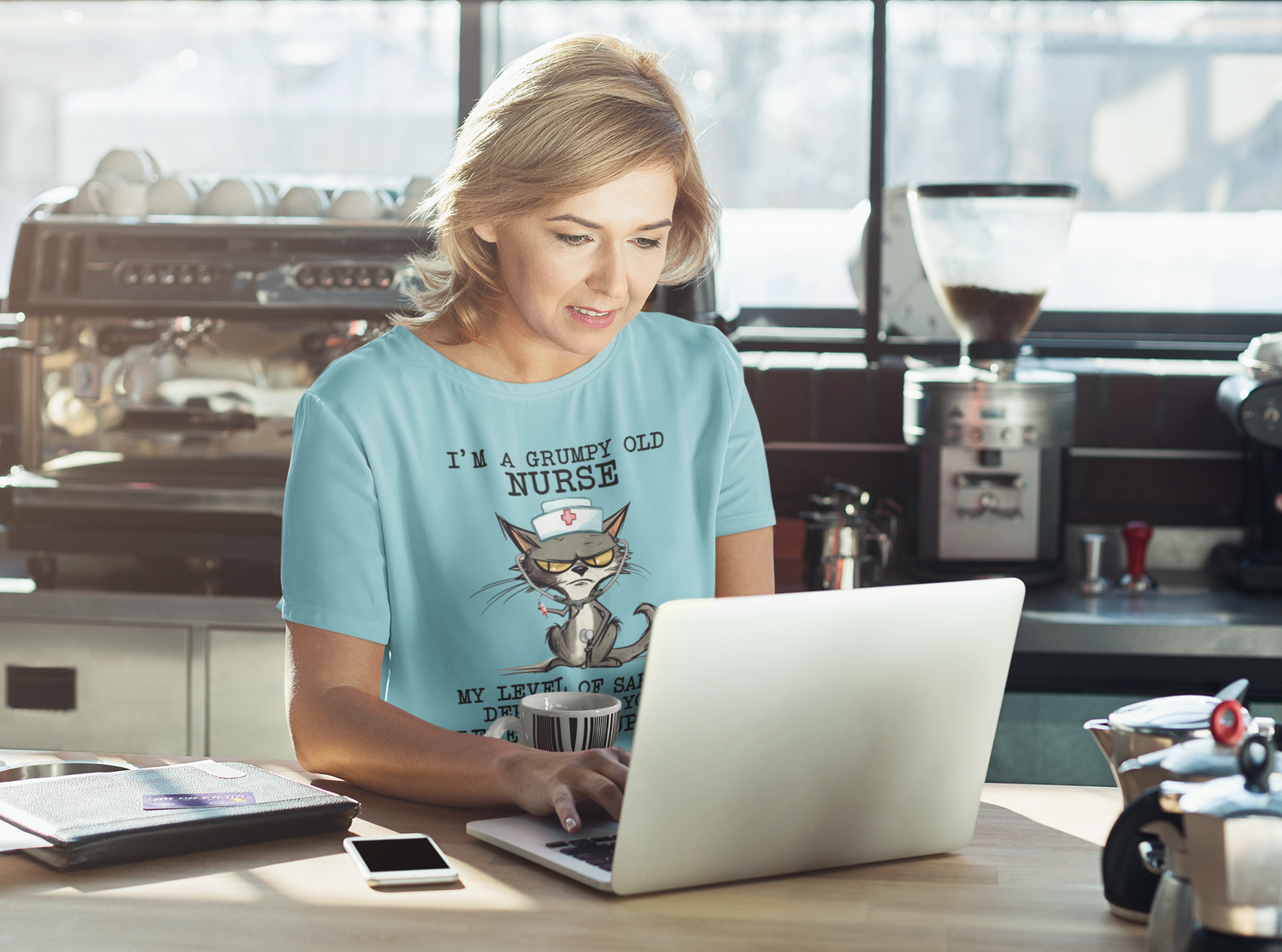Woman working on laptop at coffee shop wearing the Funny Nurse T-Shirt with grumpy cat design and witty sarcasm quote, premium turquoise cotton fabric