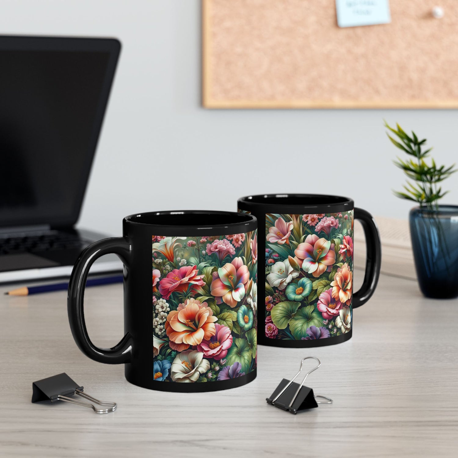 Two 11 oz black floral mugs with vivid flower designs on a modern office desk with laptop, binder clips, and a plant in the background
