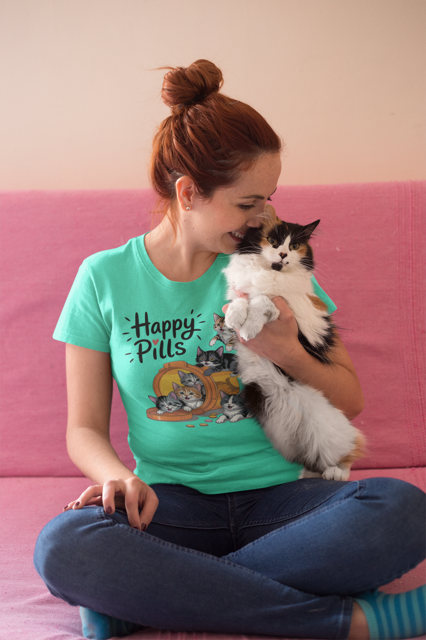 Woman in a heather sea green Happy Pills t-shirt sitting cross-legged and hugging her fluffy cat with joy